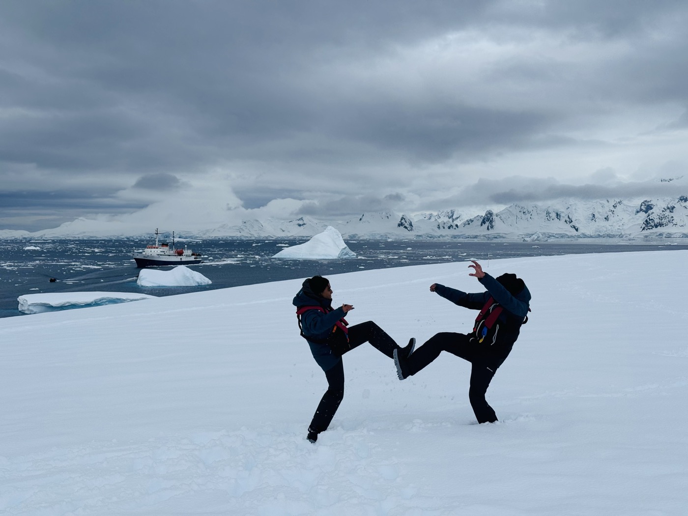 Two people play-fighting on the ice with an expedition ship and icebergs behind